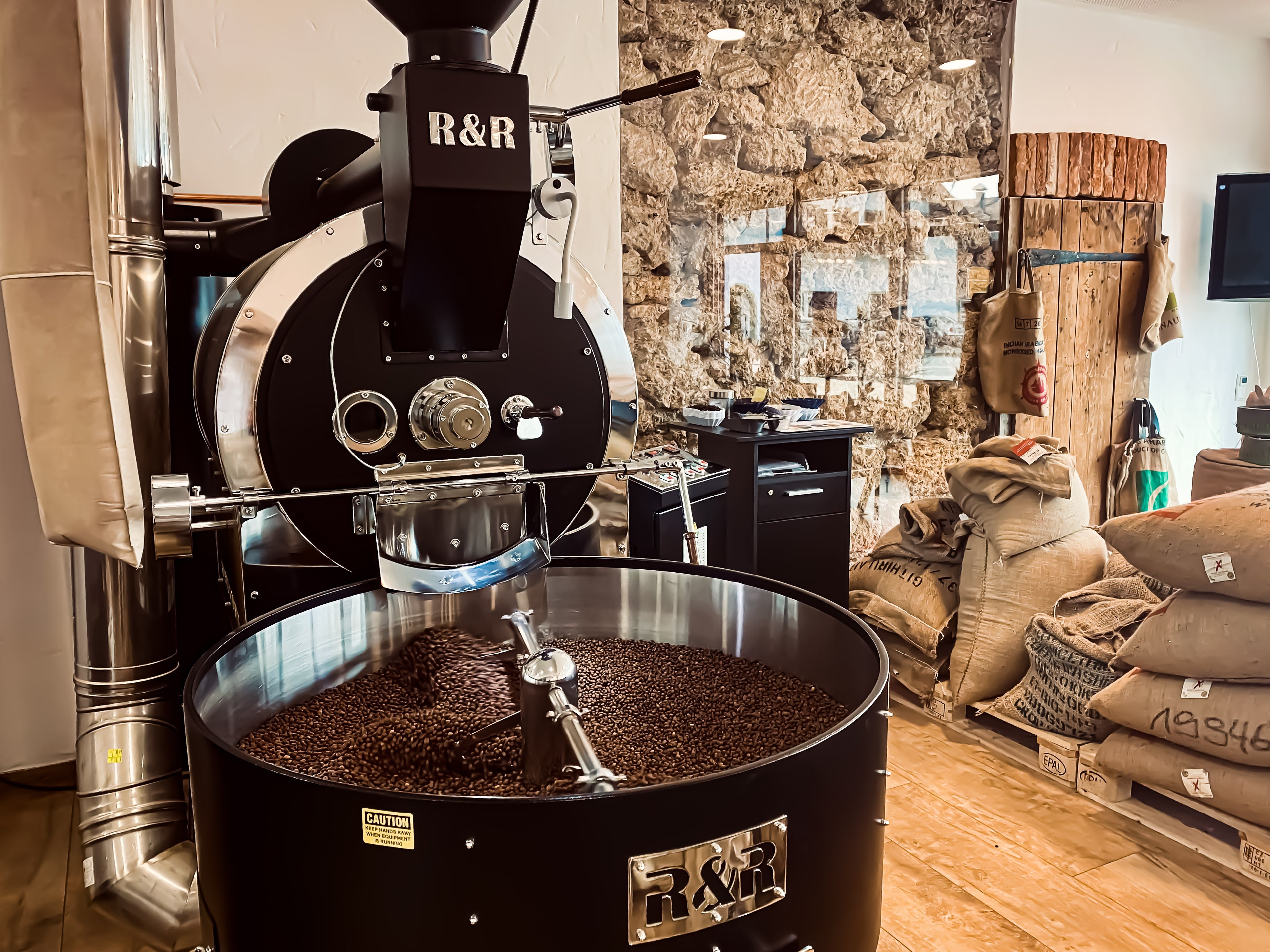 Großer Trommelröster mit frischem Kaffee in der Kühlsieb zwischen Rohkaffeesäcken. 